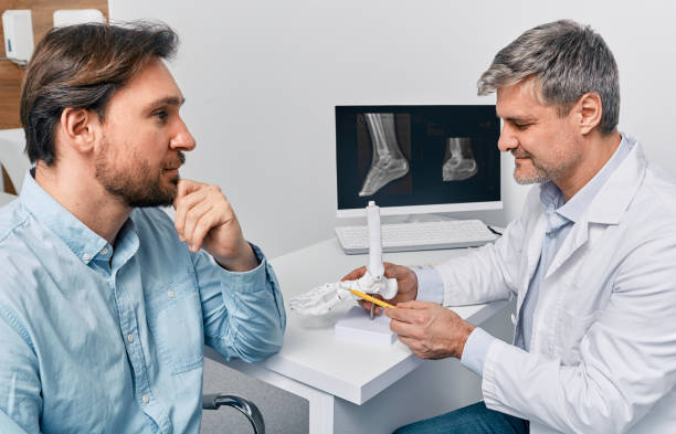Orthopedic doctor consulting adult man with leg injury and explaining treatment methods of trauma using anatomical model of foot. Treatment of trauma and fracture of leg